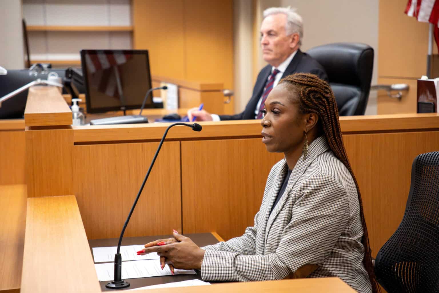 A woman at the jury stand in a courtroom, with a judge in the background.
