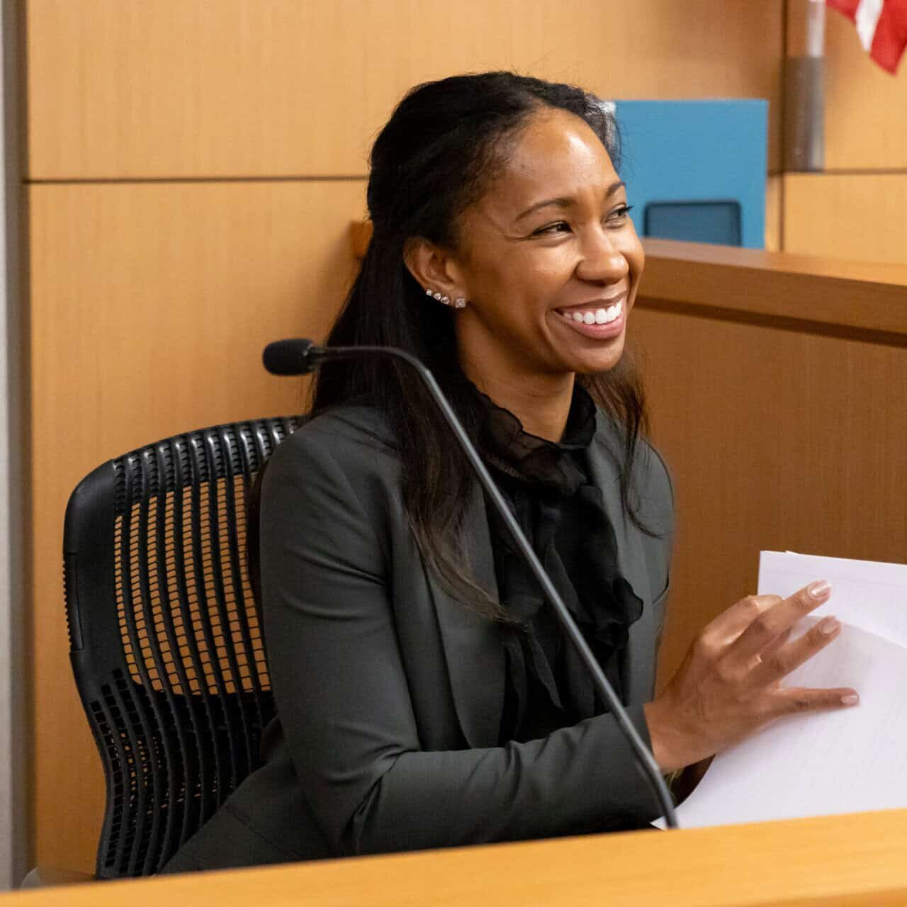 A woman smiling and holding papers at a desk in a courtroom